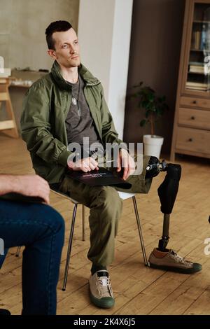 Young man with leg prosthesis sitting on chair at psychological session and describing his mental problems to counselor and other attendants Stock Photo