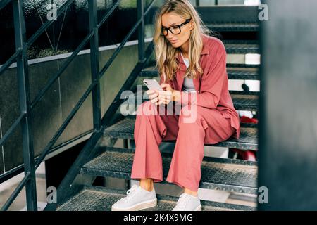 Young casual woman using smartphone while sitting on the iron steps of a modern office building. Stock Photo