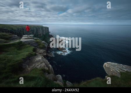 A red-jacketed hiker on top of a cliff looks out over the magnificent scenery of the sea bay. Dunnet Head, Scotland  Stock Photo
