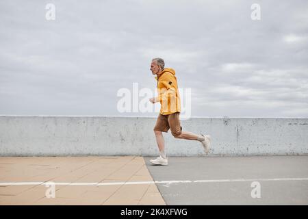 Minimal side view portrait of handsome mature man running by river against grey sky Stock Photo