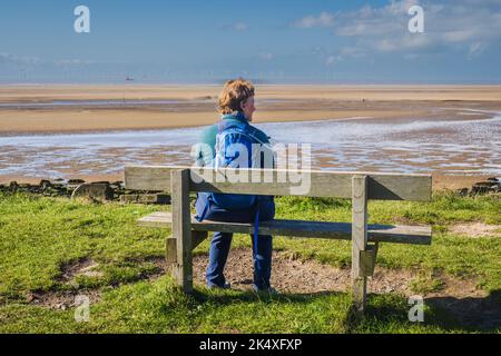 02.10.2022 Crosby, Liverpool. Merseyside, UK. Female walker wearing blue and green sat on a bench looking the Mersey estuary Stock Photo
