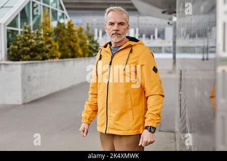Waist up portrait of active mature man looking at camera outdoors while wearing contrasted yellow jacket in city setting, copy space Stock Photo
