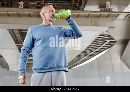 Low angle portrait of handsome senior man drinking water during outdoor workout in urban setting under bridge structure, copy space Stock Photo