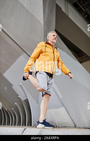 Vertical full length shot of senior man stretching legs outdoors while arming up for jogging Stock Photo