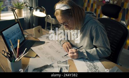 Female animator creates sketches for the cartoon. Looks at the laptop screen infront of her and draws on the paper. Comics Compilation. Storyboard movie layout for pre-production. Stock Photo