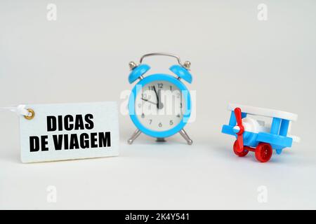 On a white surface there is a toy plane, an alarm clock and a sign with the inscription - Travel tips. Vacation concept. Text in Portuguese. Stock Photo