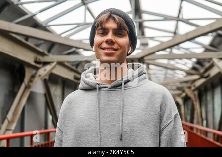 Urban portrait of smiling hipster guy wearing grey hoodie and hat with white wireless earbuds headphones. Stock Photo