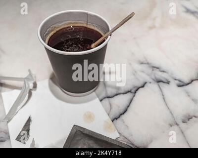 Black coffee in the brown disposable cup, a torn bag of  sugar and the wooden stirring stick on the marble table top background Stock Photo