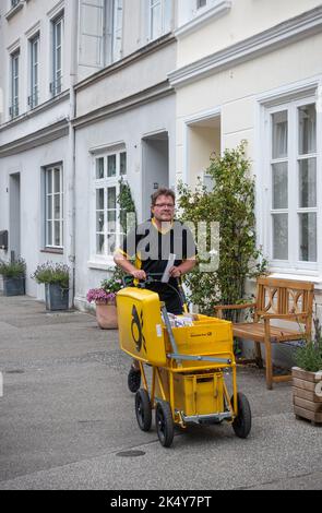 Germany, Lubeck - July 13, 2022: Closeup of Mailman pusheing yellow handcar in street in front of white painted house facades Stock Photo