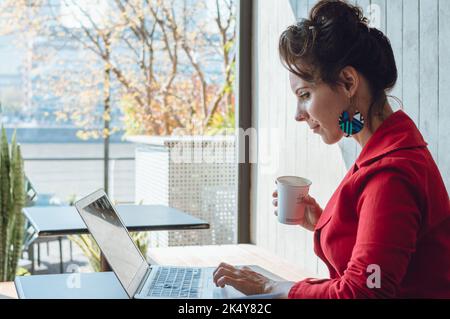 young caucasian business woman, sitting in a coffee shop with her laptop analyzing financial graphs, looking at the computer and with a cup of coffee Stock Photo