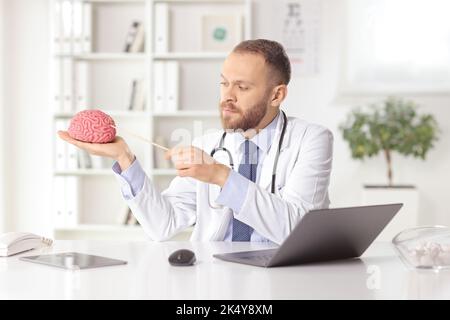 Young doctor sitting in an office and pointing at a human brain model with laptop computer on the desk Stock Photo