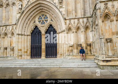 Man sitting on the carved stone seating at the entrance to York cathedral on a wet and raining afternoon, York, Yorkshire, England. Stock Photo