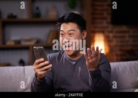 Asian Man celebrating mobile success by showing yes gesture. Excited asian man getting great news, making winner gesture and looking at phone screen Stock Photo
