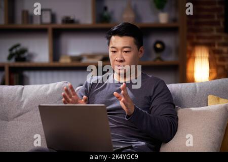 Asian man holding video conference looking at laptop webcam, working from home. Man having online meeting and communicating remotely, sitting on the Stock Photo