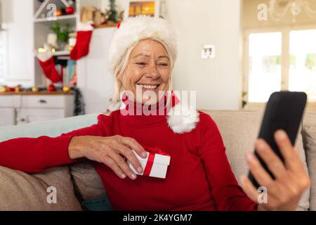 Happy senior caucasian woman wearing santa claus hat, using smartphone for video call Stock Photo