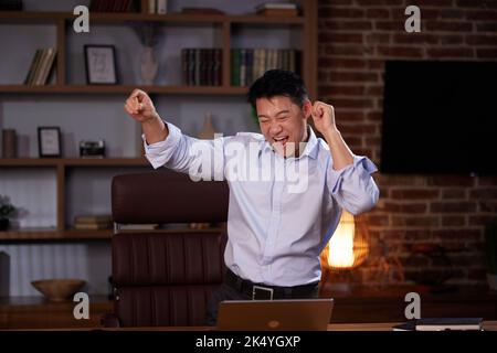 Happy Asian man enjoying music using a laptop and dancing in modern workroom. Man freelancer having fun after work, moving body near working desk at Stock Photo