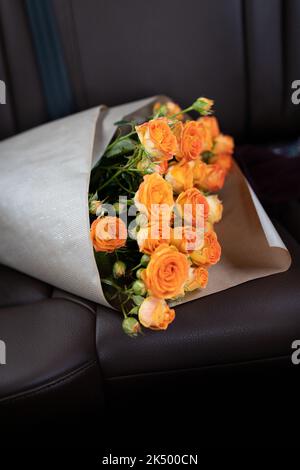 A bouquet of orange bush roses on the back seat of a car with a leather interior. Vertical photo. Stock Photo
