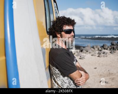 Caucasian nomad man portrait by the sea leaning on his camper van. His surfboard is standing beside. Stock Photo