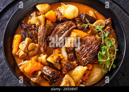 Beef Bourguignon, Beef Burgundy, stew with beef, bacon, carrots, onion bulbs and mushrooms in rich red wine sauce in black bowl on wooden table, close Stock Photo