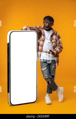 Smiling african man standing near huge smartphone with blank screen on yellow studio background Stock Photo