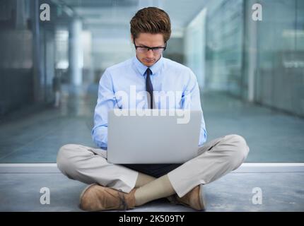 Working from anywhere. A young businessman sitting on the floor of his empty office working on a laptop. Stock Photo