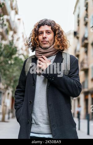 Low angle of self assured male in smart casual clothes with backpack correcting scarf and looking at camera on blurred background of city street Stock Photo