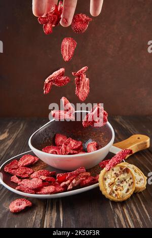 Levitation, Strawberry chips fall into ceramic plate with wooden handle and cookies with chocolate and cream on dark wooden background. Hundred Stock Photo