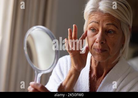 Happy senior caucasian woman sitting on bed in bedroom, looking at mirror and touching her face Stock Photo