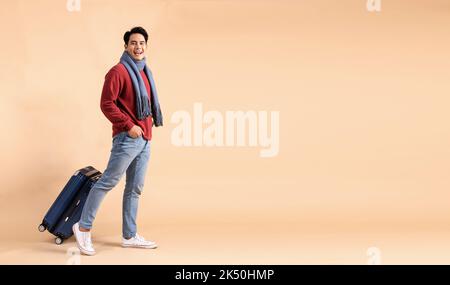 Happy Asian male tourist with luggage in red sweater and scarf enjoying winter vacation isolated on beige color background. Excited fashionable 20s guy going on a trip - Full body length studio shot. Stock Photo