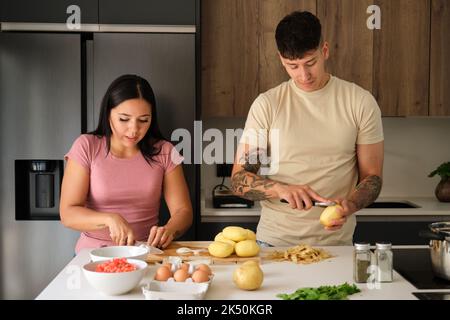 Two young latin people peeling potatoes and cutting onion at kitchen. Stock Photo