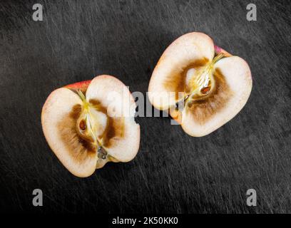 Two halves of the inside of a cut open and rotten apple, resting on top of dark cutting board. Stock Photo