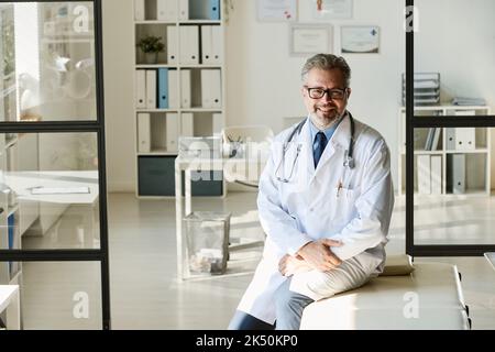 Portrait of mature doctor in white lab coat smiling at camera while working at office Stock Photo