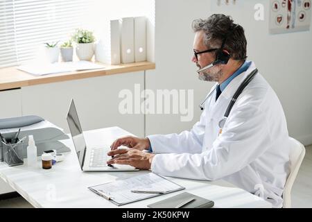 Mature doctor in headphones typing on laptop and talking to patient online at his workplace at office Stock Photo
