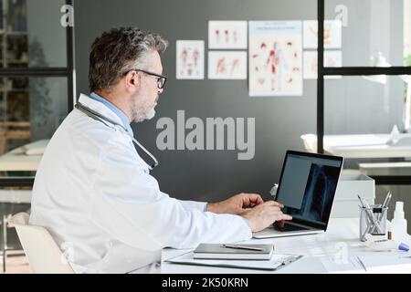 Mature medical professional examining x-ray image on laptop at table at his office Stock Photo