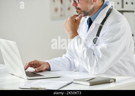 Close-up of mature general practitioner in white coat working at his workplace with laptop Stock Photo