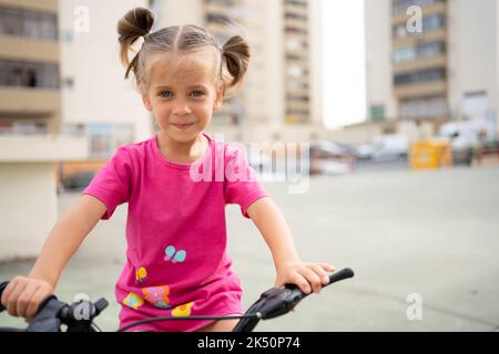 Cute little smiling girl riding bike bicycle in city on parking sunny summer day. Active family leisure with kids. Little caucasian girl 5 years old have fun with pink child bicycle Stock Photo