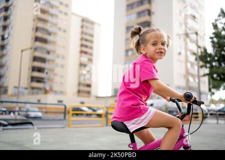 Cute little smiling girl riding bike bicycle in city on parking sunny summer day. Active family leisure with kids. Little caucasian girl 5 years old have fun with pink child bicycle Stock Photo