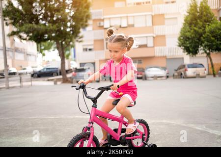 Cute little smiling girl riding bike bicycle in city on parking sunny summer day. Active family leisure with kids. Little caucasian girl 5 years old have fun with pink child bicycle Stock Photo