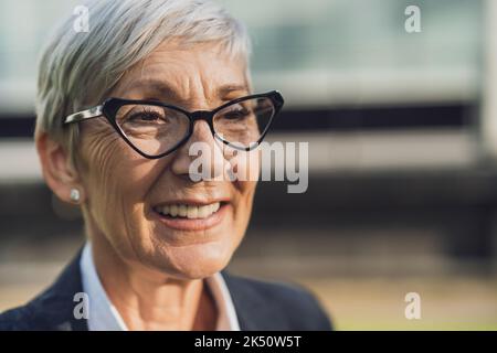 Outdoor head shot portrait of successful senior businesswoman. Happy business person is smiling and looking away. Stock Photo