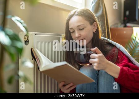 A woman wrapped in a plaid is sitting on the floor with hot tea and warming herself near a warm radiator. The concept of economy and the crisis of ene Stock Photo