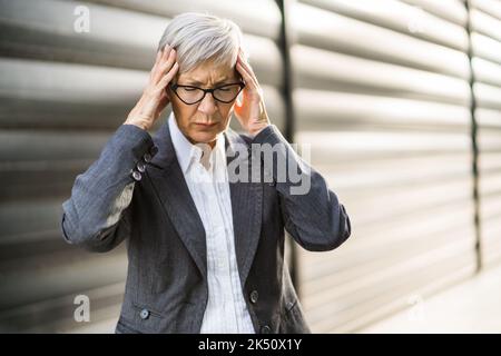 Senior businesswoman is having headache. She is standing in front of company building. Stock Photo