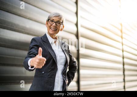 Outdoor portrait of successful senior businesswoman in front of company building. She is looking at camera and showing thumbs up. Stock Photo