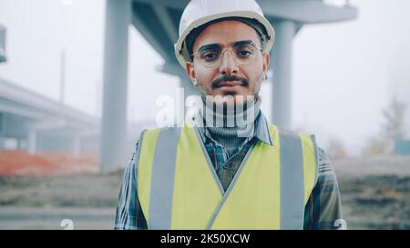 Portrait of Middle Eastern builder wearing uniform standing outdoors in building site and looking at camera. Profession and specialists concept. Stock Photo