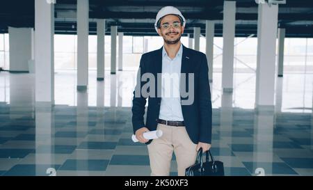 Portrait of young ambitious Middle Eastern engineer standing in new empty building holding blueprint and briefcase and looking at camera Stock Photo