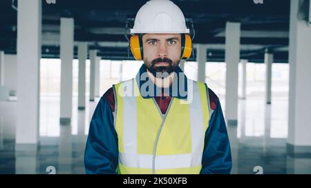 portrait of construction worker wearing safety helmet and headphones indoors in new empty industrial building. People and profession concept. Stock Photo