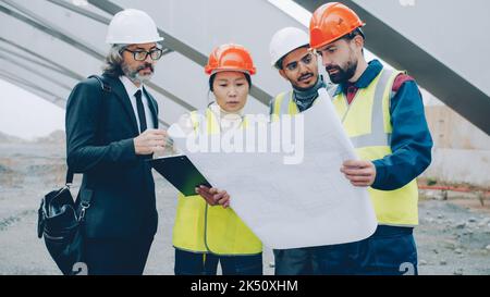 Construction workers are discussing project with manager standing in building area reading papers and looking around. People and occupation concept. Stock Photo