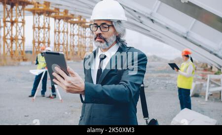 Architect making online video call with tablet talking and looking at display standing at construction site while builders are working in background Stock Photo