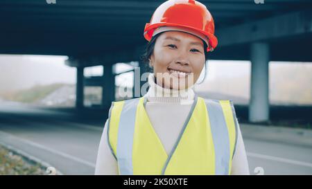 Asian forewoman wearing vest and helmet is standing outside in construction site smiling enjoying building career. People and occupation concept. Stock Photo