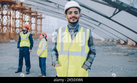 portrait of Arab man wearing safety helmet standing in construction site smiling looking at camera while people in uniform working in background Stock Photo