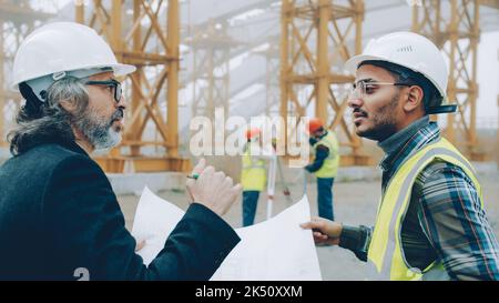 Foreman is discussing blueprints with project manager while surveyors are working in background. Construction site and workers concept. Stock Photo